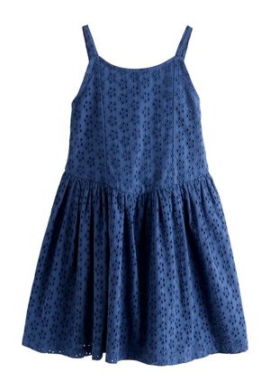 Niña con cabello castaño claro que lleva un vestido sin mangas azul marino, de pie en una calle cerca de una pared blanca y una tubería roja.