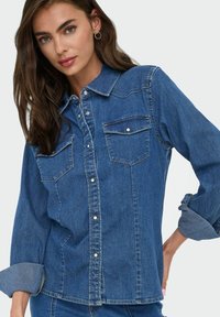 Keskmise sinise värvi denimpait, millel on nööpidega esiosa, kaks rinna taskut ja üles keritud varrukad, millel on kontrastsetest denimmaterjalist mansetid.
