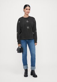 Kvinne iført en svart genser med stjernemønster, blå oppbrettede jeans, svarte platåboots, og holder en liten svart håndveske mot en ensfarget bakgrunn.