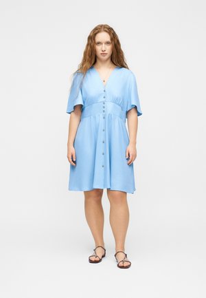 Mujer con cabello largo y rojo lleva un vestido azul claro abotonado y sandalias negras, de pie frente a un fondo blanco liso.