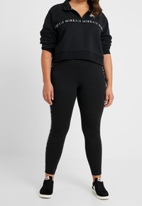 Svart kort sweatshirt med dragkedja i kragen och logotyp, ihop med svarta leggings med sidobranding. Bärs med svarta sneakers.