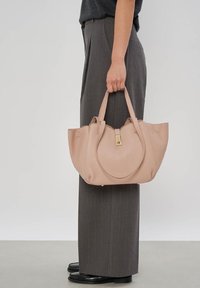 Personne portant un pantalon gris à jambes larges et des chaussures noires tient un grand sac à main en cuir beige avec des ferrures dorées par ses poignées.