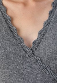 Zbliżenie osoby w szarym, dzianinowym swetrze z wykrojem w serek w kształcie falbany, ukazujące górną część klatki piersiowej i detale dekoltu.