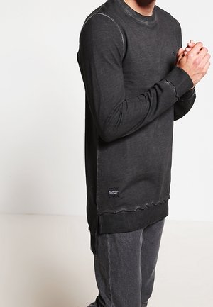 Mężczyzna ubrany w czarną, długorękawową bluzę z postrzępionym dołem oraz szare spodnie, stojący z rękami złączonymi na tle o jednolitym kolorze.