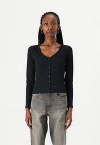 Svart ribbet cardigan med V-hals, lange ermer og seks frontknapper, kombinert med lysegrå løstsittende jeans.