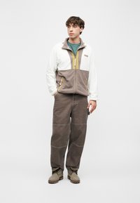 Νέος άνδρας στέκεται με τα χέρια στις τσέπες φορώντας φλις μπουφάν με χρωματικούς συνδυασμούς, καφέ παντελόνι, πράσινο πουκάμισο και μπεζ μπότες σε λευκό φόντο.