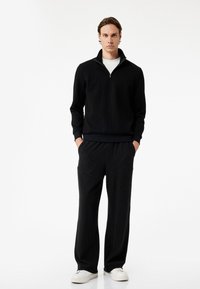 Sort lynlås sweater med ribbet tekstur, parret med løstsiddende sorte stribede bukser og hvide sneakers. Simpelt design.