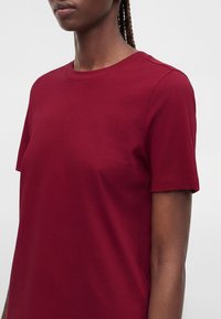 Femme portant un T-shirt bordeaux uni à manches courtes avec un col rond, visible du cou jusqu'au milieu du torse sur un fond uni.