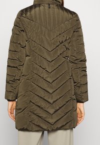 Rückansicht einer Person, die eine olivgrüne gesteppte Winterjacke mit Zickzacknähten und beigen Hosen vor einem einfarbigen Hintergrund trägt.