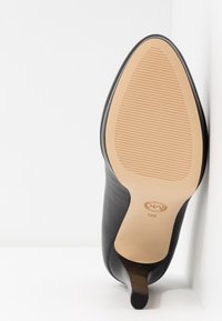 Chaussure en cuir noir à bout pointu avec semelle beige montrant une adhérence texturée et le marquage de taille 8M, positionnée verticalement sur un fond blanc.