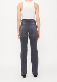Personne portant un jean noir délavé taille haute et coupe droite et un haut blanc sans manches, debout de dos sur un fond uni.