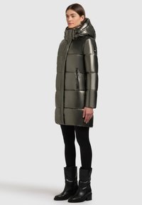Gevoerde parka in glanzend olijfgroen met een hoge kraag, een ritssluiting aan de voorkant en twee ritszakken. Gecombineerd met zwarte legging en kniehoge laarzen.