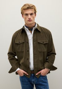 Olivgrüne Cordjacke mit zwei Brusttaschen, Knopfverschluss und Kentkragen, über einem weißen Hemd und blauen Jeans getragen.