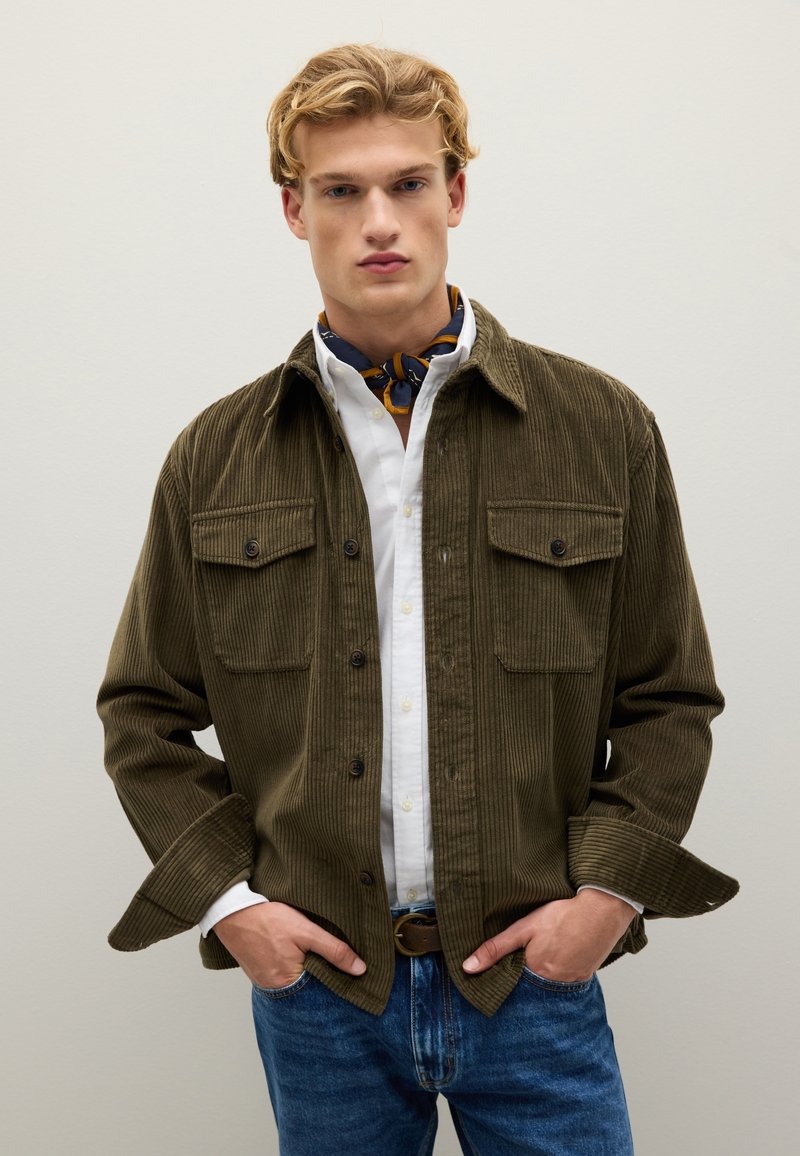 Olivgrüne Cordjacke mit zwei Brusttaschen, Knopfverschluss und Kentkragen, über einem weißen Hemd und blauen Jeans getragen.