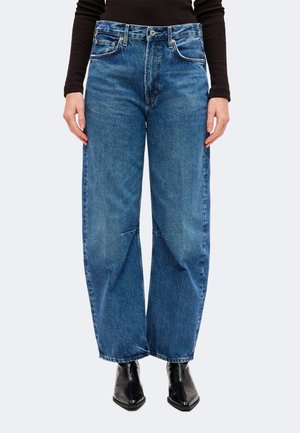 Persona que lleva vaqueros azules de mezclilla sueltos con cintura alta, combinados con botas negras de punta afilada y un top negro de manga larga.