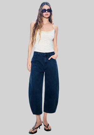 Top bianco senza maniche con piccole decorazioni, abbinato a jeans a gamba larga blu navy e sandali neri con tacco. Il modello si trova davanti a uno sfondo grigio.