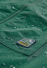 Tissu imperméable vert avec des gouttes d'eau, mettant en valeur un patch cousu avec un motif de crocodile et le texte "Next 82".