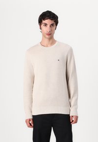 Pull en tricot beige avec un col rond, des manches longues et un petit logo brodé sur la poitrine, associé à un pantalon sombre.