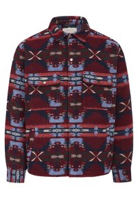 Veste chemise à carreaux rouges et bleus avec deux poches avant, ornée de motifs géométriques et de pressions métalliques, fabriquée à partir d'un tissu texturé.