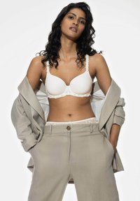 Sostén blanco acolchado con ribete floral, combinado con pantalones cortos en beige claro. La modelo lleva un blazzer abierto a juego sobre el conjunto.