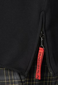 Tissu noir avec une fermeture éclair noire partiellement ouverte révélant une languette rouge étiquetée "HUGO BERLIN" sur un tissu à carreaux gris.