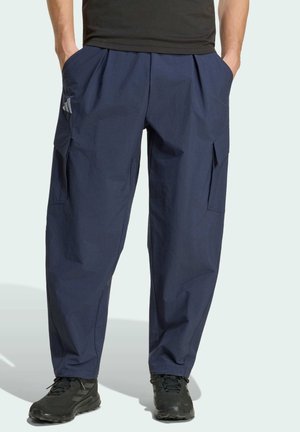 Mężczyzna ubrany w granatowe spodnie typu cargo z bocznymi kieszeniami i czarne trampki, ręce włożone do kieszeni, stojący na jednolitym tle.