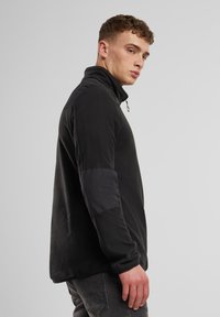 Svart genser med høy krage og halv glidelås, med kontrasterende teksturert stoff på skuldrene og ermene.