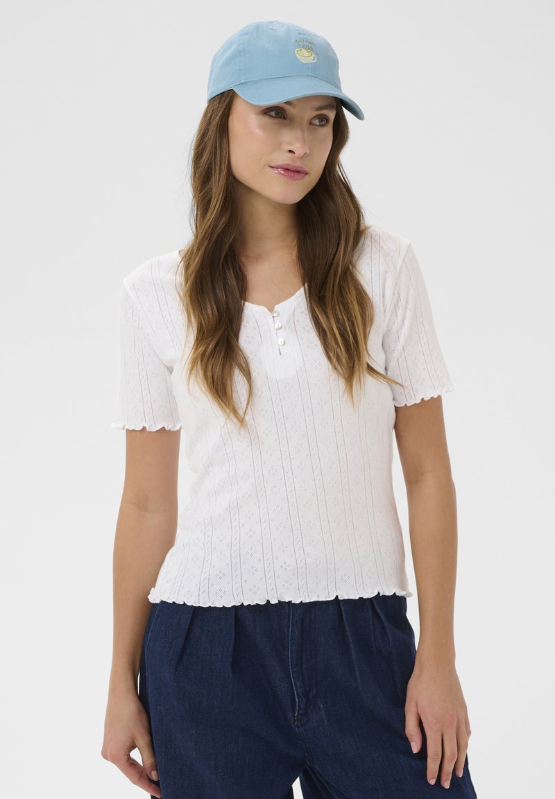 Femme portant une casquette bleu clair avec un motif de citron, un haut blanc à manches courtes boutonné et un pantalon bleu foncé, debout devant un fond uni.