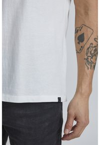 Hvit bomulls t-skjorte med rett nederkant og svart etikett på siden, kombinert med mørke jeans. Synlige tatoveringer på underarmen.