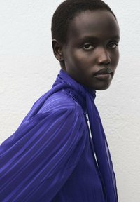 Blusa viola con righe verticali, caratterizzata da un collo alto e dettaglio di sciarpa annodata, realizzata in un tessuto leggero e trasparente.