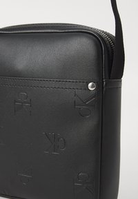 Bolso cruzado sintético negro con patrón de logo en relieve, detalles en hardware plateado y acabado texturizado, con cierre de cremallera y correa ajustable.
