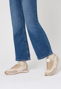 Baskets de sport beiges avec un suède texturé et des matériaux synthétiques, à lacets, et une semelle en caoutchouc blanche, associées à un pantalon en denim bleu cropped.