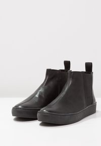 Botas de tobillo de cuero negro con un acabado liso, paneles laterales elásticos, lengüetas para tirar y una suela gruesa negra. Diseño simple y aerodinámico.