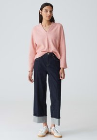 Hellrosa V-Ausschnitt-Pullover, lockere Passform; dunkle Denim-Hosen mit weitem Bein und umgeschlagenem Saum; weiße Sneaker mit pastellfarbenen Akzenten.