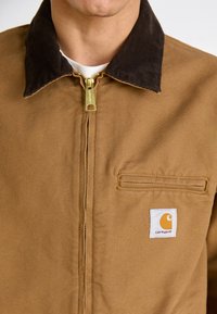 Chaqueta acolchada marrón con cuello de pana oscuro, cierre frontal con cremallera, bolsillo de parche y logo de Carhartt en naranja sobre un fondo blanco.