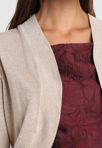Beige cardigan with a soft texture, draped open front, and wide collar, layered over a burgundy dress with a floral pattern.