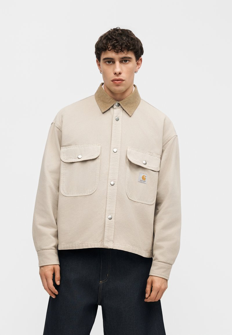 Joven con cabello corto y rizado, vistiendo una chaqueta beige con botones, cuello marrón claro y bolsillos con solapa, combinada con pantalones oscuros, mirando hacia adelante.