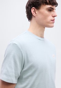 Νεαρός άνδρας που φοράει ανοιχτό γαλάζιο υφασμάτινο πουκάμισο με κοντά μανίκια, κοιτάζοντας προς τα δεξιά, με φόντο απλό ανοιχτόχρωμο.