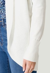 Cardigan en tricot blanc avec texture côtelée, devant ouvert et manches longues. Associé à un jean bleu, mettant en valeur un look décontracté et superposé.