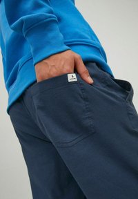 Marineblauwe broek met een gladde textuur, voorzien van een achterzak en een klein wit merklabel zichtbaar op de zak. Gecombineerd met een blauwe sweatshirt.