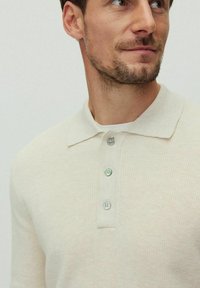 Vaalean kermanvärinen pitkähihaista polo paitaa, teksturoitu kangas, pehmeä kaulus, kolme nappia kauluksessa sinivihreillä yksityiskohdilla, rento muotoilu.