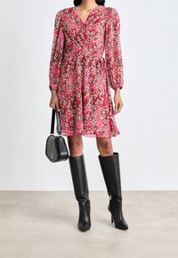Femme portant une robe florale rouge, des bottes noires à talons hauts jusqu'aux genoux, et tenant un sac à main noir devant un fond uni.