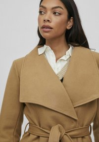 Abrigo de lana color camel, con cinturón, cuello ancho y diseño envolvente. Llevado sobre una camisa blanca y un chaleco de punto con escote en V.