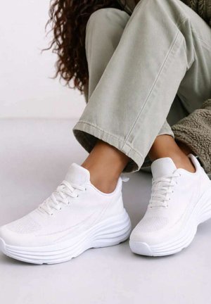 Personne portant un pantalon beige et des baskets blanches épaisses, assise jambes croisées sur un sol clair.