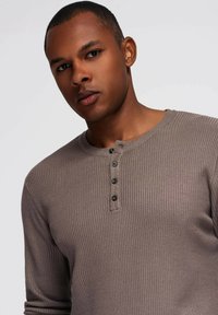 Camisa henley de manga larga marrón con canalé, que presenta un escote redondo y cinco botones negros en la parte frontal. Textura suave y diseño ajustado.