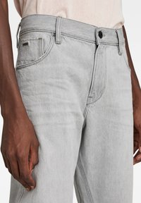 Los jeans de mezclilla gris claro presentan un diseño clásico de cinco bolsillos, costuras visibles y un cierre de botón metálico en la cintura.