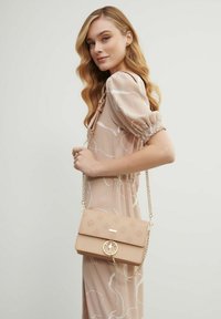 Kobieta z długimi falującymi blond włosami, ubrana w beżową sukienkę w wzory, trzymająca beżową torebkę z złotym łańcuchem na ramieniu.