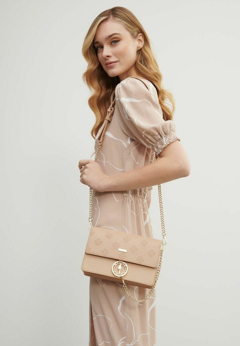 Kobieta z długimi falującymi blond włosami, ubrana w beżową sukienkę w wzory, trzymająca beżową torebkę z złotym łańcuchem na ramieniu.