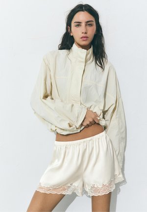 Vrouw met lang donker haar draagt een losse crèmekleurige jas en zijdezachte crèmekleurige shorts met kanten randje, staand tegen een effen lichte achtergrond.