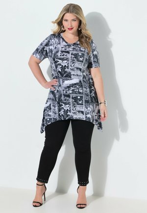 Vrouw poseert in zwart-wit tuniek met korte mouwen, zwarte leggings met studs en zwarte sandalen met hoge hakken tegen een witte achtergrond.